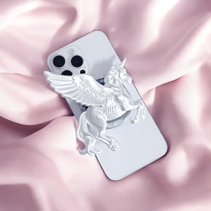 Può includere: Un telefono bianco con un supporto per telefono a forma di unicorno alato scolpito. Il supporto è attaccato al retro del telefono. Il telefono e il supporto sono appoggiati su uno sfondo rosa e serico.