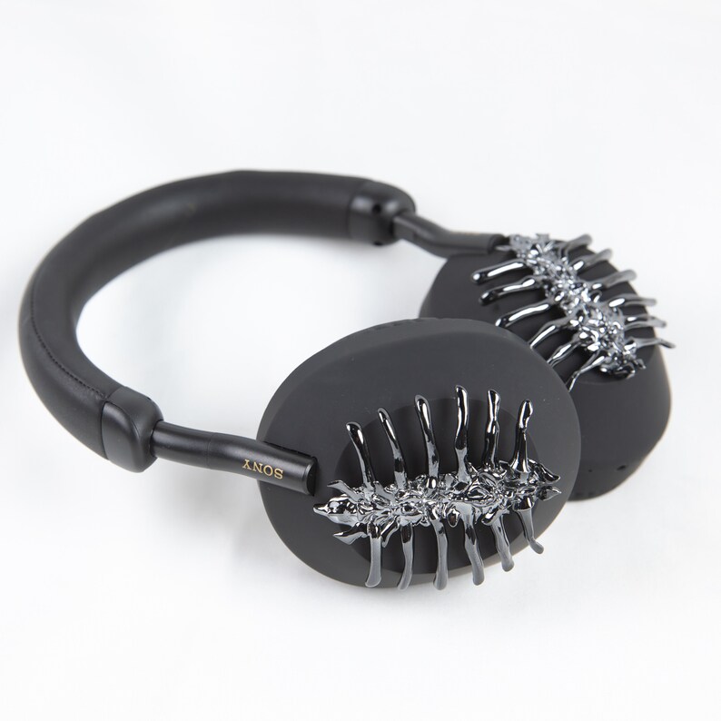 May include: Black over-ear headphones with a unique design featuring a silver, skeletal-like pattern on each earcup. The headphones have a black leather headband and a gold "SONY" logo on the side.