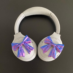 May include: White wireless headphones with a blue and purple iridescent bow on each earcup.