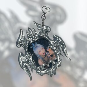 May include: A silver-colored keychain with a heart-shaped clasp and a decorative frame. The frame has an ornate, gothic design with wing-like elements and holds a small photo of a person with blue hair.