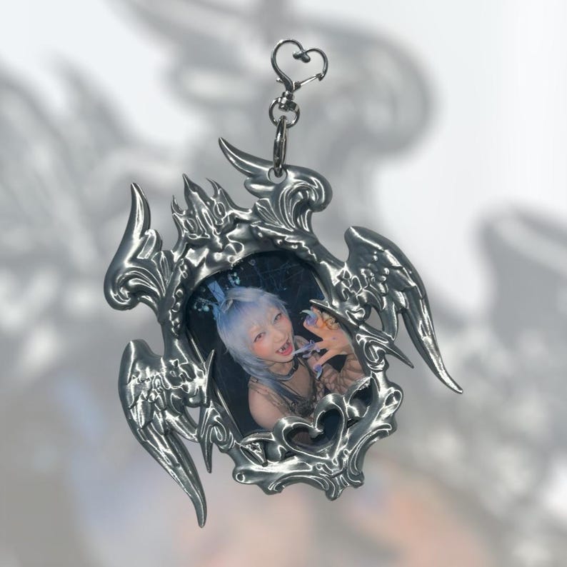 May include: A silver-colored keychain with a heart-shaped clasp and a decorative frame. The frame has an ornate, gothic design with wing-like elements and holds a small photo of a person with blue hair.