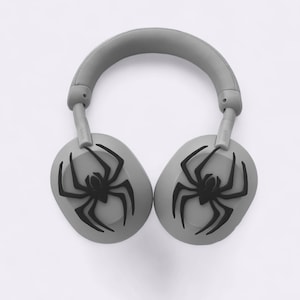 Può includere: Cuffie grigie over-ear con un disegno di ragno nero su ciascun padiglione auricolare. L'archetto e i padiglioni sono grigi, con un disegno di ragno nero. Progettate per l'ascolto audio.
