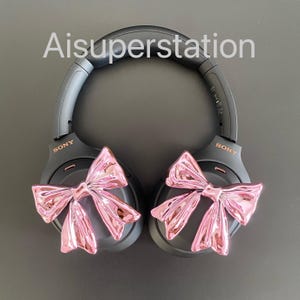 Pu&ograve; includere: Cuffie over-ear nere con fiocchi rosa attaccati a ciascun padiglione auricolare. Le cuffie hanno la scritta "SONY" stampata su ciascun padiglione auricolare.
