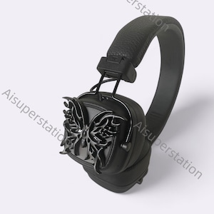 Può includere: Cuffie over-ear nere con un design a farfalla sul padiglione auricolare. Le cuffie hanno un archetto e padiglioni imbottiti. Il design a farfalla è un ornamento scuro e ornato. Le cuffie sono leggermente angolate.