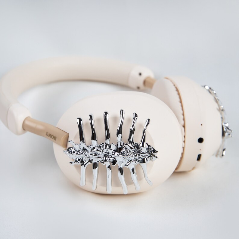 May include: Cream-colored wireless headphones with silver decorative accents. The headphones have a padded headband and earcups. The left earcup features a silver, stylized design. The brand name "SONY" is visible on the left side.