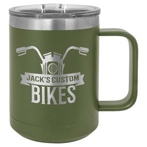 May include: An olive green travel mug with a silver rim and lid. The mug features a motorcycle handlebar graphic and the text "JACK'S CUSTOM BIKES" in white. The mug has a large, curved handle.