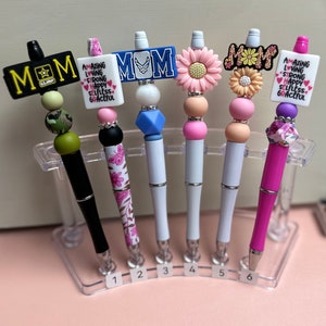 May include: Six decorative pens with various designs. The pens feature embellishments such as flowers, beads, and text that reads "MOM" and "AMAZING LOVING STRONG HAPPY SELFLESS GRACEFUL." The pens are displayed in a clear plastic holder.