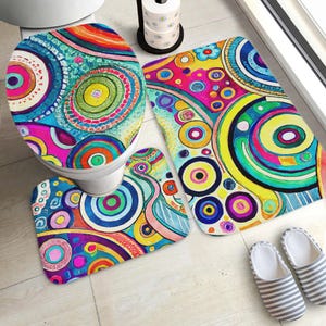 Puede incluir: Un colorido juego de baño con una funda para el asiento del inodoro y dos alfombras a juego. El juego tiene un diseño abstracto vibrante con círculos concéntricos en tonos de azul, amarillo, rosa y verde. Las alfombras son rectangulares y ovaladas.