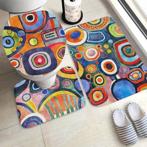 Peut inclure: Un ensemble de salle de bain comprenant une housse d'abattant de toilettes, un tapis de bain rectangulaire et un tapis de contour, ornés d'un motif abstrait multicolore composé de cercles concentriques et de formes géométriques vives.