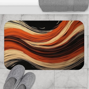 May include: A rectangular bath mat with an abstract design of flowing orange, black, and cream-colored waves. The mat is placed on a tiled floor, with a pair of gray slippers and a gray towel visible.