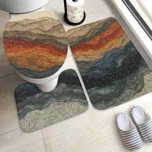 Puede incluir: Un juego de baño que incluye una funda para el asiento del inodoro, una alfombra contorneada y una alfombra de baño rectangular. El juego tiene un diseño abstracto en capas en tonos marrón, naranja y azul. Un par de zapatillas a rayas está al lado.