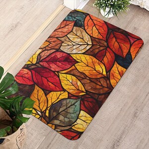 May include: A rectangular floor mat featuring a vibrant, stained-glass style pattern of overlapping autumn leaves in shades of deep red, burnt orange, golden yellow, and dark brown, with distinct black outlines defining each leaf shape.