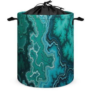 May include: A cylindrical laundry hamper with a teal and turquoise abstract pattern. The hamper has black handles and a black drawstring closure at the top. The design resembles a geode or agate stone.