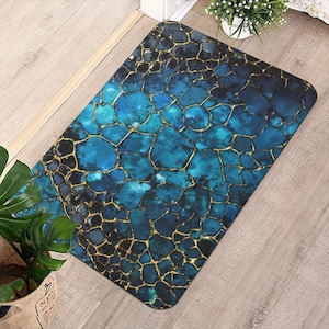 Op de afbeelding: Rechthoekige vloermat met een mozaïekontwerp in tinten blauw en zwart, geaccentueerd met goudkleurige lijnen. De mat heeft een textuur en ligt op een houten vloer.
