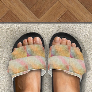 Puede incluir: Sandalias deslizantes negras con una correa estampada colorida. La correa presenta un diseño con cuadrados rosas, amarillos y naranjas. Las sandalias están sobre una alfombra de color claro, con un suelo de madera en el fondo. Calzado informal.