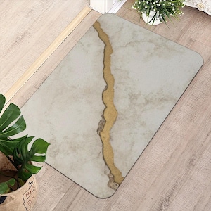 Puede incluir: Alfombra rectangular con un patrón gris claro similar al mármol y una franja central dorada irregular. La alfombra está colocada sobre un suelo de madera, con una planta en maceta.