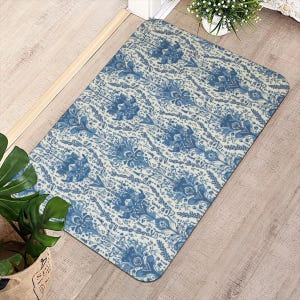 May include: Rectangular doormat with a blue and white floral pattern. The design features stylized flowers and foliage on a cream background. The mat is placed on a light wood floor, suggesting an entryway or indoor space.