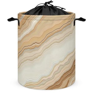 May include: A cylindrical laundry hamper featuring a beige, cream, and brown marbled agate pattern. The hamper has two black side handles and a black fabric drawstring closure at the top, providing a secure opening for the interior storage space.