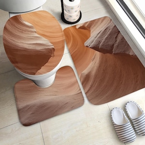 Op de afbeelding: Een driedelige badkamerset met een canyonlandschapontwerp. De set bevat een toiletbril, een contourtapijt en een rechthoekige badmat. Het kleurenpalet bestaat uit warme bruin- en beigetinten, wat een natuurlijke uitstraling creëert.