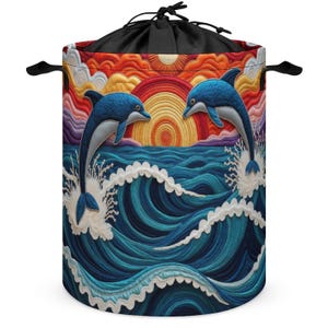 May include: A cylindrical laundry hamper featuring a quilted-style graphic of two blue dolphins leaping from deep blue ocean waves towards a vibrant orange and red sunset. The hamper has a black drawstring closure at the top and two side handles.