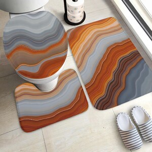 Può includere: Un set da bagno composto da tre pezzi: un copriwater, un tappetino da bagno rettangolare e un tappetino sagomato a U. Ogni elemento presenta un motivo ondulato ispirato all'agata nei toni dell'arancione, del terracotta, del grigio e del bianco.