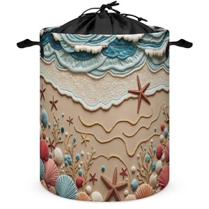 Könnte beinhalten: Ein zylindrischer Wäschekorb mit einem Strandmotiv, das blaue Meereswellen, weißen Schaum und einen sandigen Untergrund zeigt. Das Design umfasst verstreute Seesterne, verschiedene Muscheln in Rosa, Blau und Weiß sowie korallenartige Texturen.
