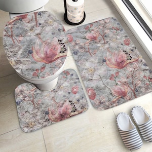 May include: A three-piece bathroom rug set with a floral design. The set includes a toilet seat cover, a contour rug, and a larger bath mat. The rugs feature a watercolor-style floral pattern on a gray background.