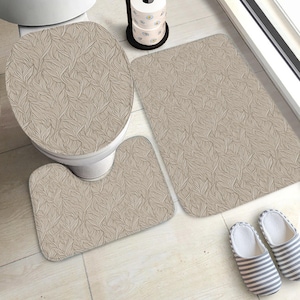 Op de afbeelding: Een beige badkamerset met een bladerenpatroon, bestaande uit een toiletbril, een contourtapijt en een rechthoekige badmat. De set wordt gecompleteerd door gestreepte pantoffels. Een rol toiletpapier is ook zichtbaar.