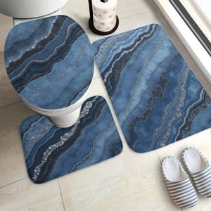 Pode incluir: Um conjunto de casa de banho azul e prateado com uma capa para o assento da sanita, um tapete de contorno e um tapete de banho. O conjunto tem um design marmoreado com tons de azul e prata. Um par de chinelos listrados está no chão.