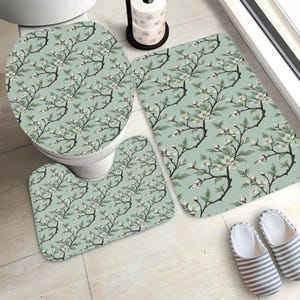 Könnte beinhalten: Ein dreiteiliges Badezimmer-Set mit floralem Muster. Das Set beinhaltet einen Toilettensitzbezug, eine Konturmatte und eine rechteckige Badematte. Das Design zeigt weiße Blüten und grüne Blätter auf hellgrünem Hintergrund.