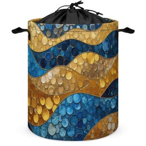 Puede incluir: Una cesta de lavandería cilíndrica con cierre de cordón y asas. El exterior presenta un patrón repetido de círculos azules y dorados dispuestos en bandas onduladas. La parte superior y las asas son negras.