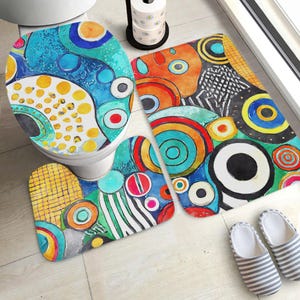 May include: A bathroom set featuring a toilet seat cover, a rectangular bath mat, and a U-shaped contour rug, all decorated with a vibrant, multi-coloured abstract pattern of circles, dots, and geometric shapes in blue, orange, yellow, and black.