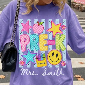 Pode incluir: Uma t-shirt violeta com uma impress&atilde;o gr&aacute;fica contendo o texto PRE-K e Mrs. Smith. O design inclui uma ma&ccedil;&atilde; xadrez, estrelas, um rosto sorridente e uma caixa de l&aacute;pis de cor com a etiqueta Teach sobre riscas azuis verticais e pontos.