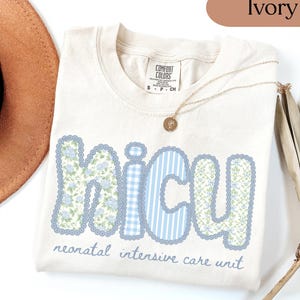 May include: Ivory-colored t-shirt with the word "NICU" in a blue floral and striped design. The text "neonatal intensive care unit" is below the word. A gold necklace with a pendant is on the shirt.