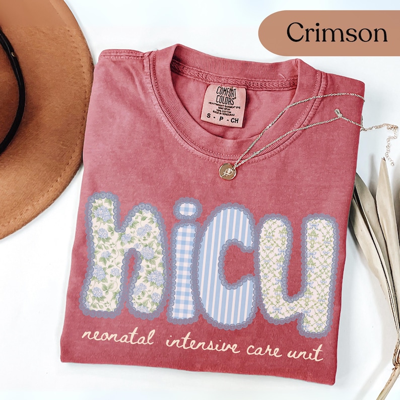 May include: Crimson-colored t-shirt with the letters "NICU" in a patchwork design. The letters are decorated with floral and striped patterns. The text "neonatal intensive care unit" is below the letters. A gold necklace with a pendant is on the shirt.