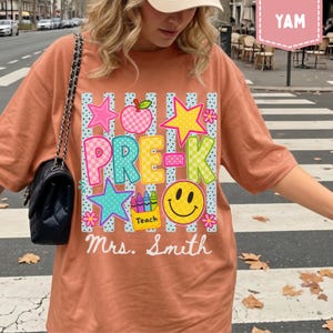 Pode incluir: Uma t-shirt cor de terracota com uma estampa gr&aacute;fica contendo o texto PRE-K e Mrs. Smith. O design inclui uma ma&ccedil;&atilde; xadrez, estrelas, um rosto sorridente e uma caixa de giz de cera com a etiqueta Teach sobre listras verticais azuis.