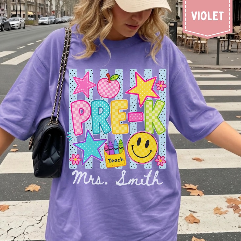 Pode incluir: Uma t-shirt violeta com uma impress&atilde;o gr&aacute;fica contendo o texto PRE-K e Mrs. Smith. O design inclui uma ma&ccedil;&atilde; xadrez, estrelas, um rosto sorridente e uma caixa de l&aacute;pis de cor com a etiqueta Teach sobre riscas azuis verticais e pontos.