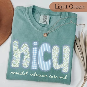 May include: Light green t-shirt with the word "NICU" in a floral and striped design. The text "neonatal intensive care unit" is below the word "NICU". A silver necklace with a pendant is on the shirt.