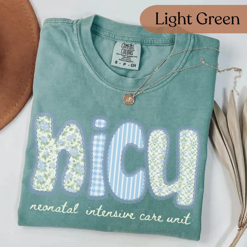 May include: Light green t-shirt with the word "NICU" in a floral and striped design. The text "neonatal intensive care unit" is below the word "NICU". A silver necklace with a pendant is on the shirt.