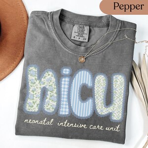 May include: A gray Comfort Colors t-shirt with the letters "NICU" in blue and white floral, gingham, and striped patterns. The words "neonatal intensive care unit" are below the letters. A silver necklace with a small pendant is on the shirt.