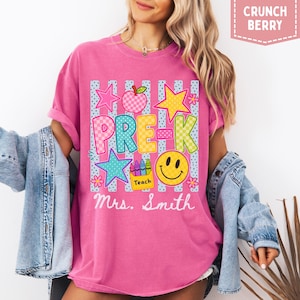 Pode incluir: Uma t-shirt rosa com um gr&aacute;fico contendo o texto PRE-K e Mrs. Smith. O design inclui uma ma&ccedil;&atilde; xadrez, estrelas, um rosto sorridente e uma caixa de l&aacute;pis de cor com a etiqueta Teach, sobre um fundo de riscas verticais azuis e brancas.