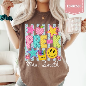 Pode incluir: Uma t-shirt castanha com um gr&aacute;fico contendo o texto PRE-K e Mrs. Smith. O design inclui uma ma&ccedil;&atilde; axadrezada, estrelas, uma cara sorridente e uma caixa de l&aacute;pis de cor com a etiqueta Teach sobre riscas verticais azuis e brancas.