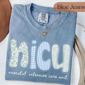May include: A blue Comfort Colors t-shirt with the word "NICU" in a floral and striped design. The text "neonatal intensive care unit" is below the letters. A gold necklace with a pendant is on the shirt.