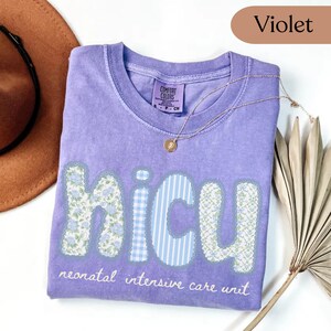 May include: Violet-colored t-shirt with the letters "NICU" in a patchwork design. The letters are decorated with floral and striped patterns. The words "neonatal intensive care unit" are printed below the letters. A gold necklace is draped on the shirt.