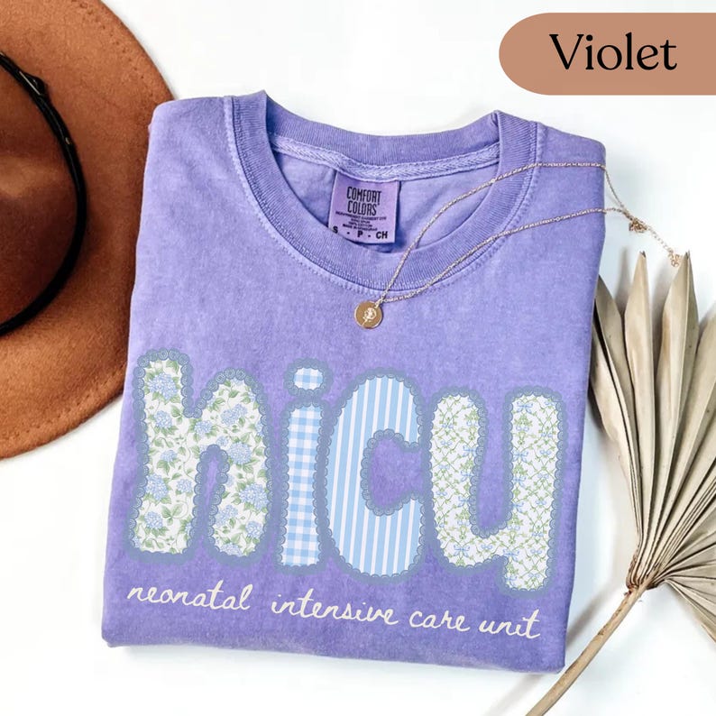 May include: Violet-colored t-shirt with the letters "NICU" in a patchwork design. The letters are decorated with floral and striped patterns. The words "neonatal intensive care unit" are printed below the letters. A gold necklace is draped on the shirt.