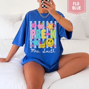 Pode incluir: Uma t-shirt azul com uma estampa gr&aacute;fica contendo o texto PRE-K e Mrs. Smith. O design inclui um rosto sorridente amarelo, uma ma&ccedil;&atilde; rosa, estrelas coloridas e uma caixa de l&aacute;pis de cor com a etiqueta Teach sobre listras verticais.