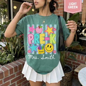 Pode incluir: Uma t-shirt de manga curta verde s&aacute;lvia com uma estampa gr&aacute;fica colorida com o texto "PRE-K" e "Mrs. Smith". O design inclui uma ma&ccedil;&atilde; rosa, estrelas amarelas e azuis, um rosto sorridente amarelo e uma caixa de l&aacute;pis com a palavra "Teach".