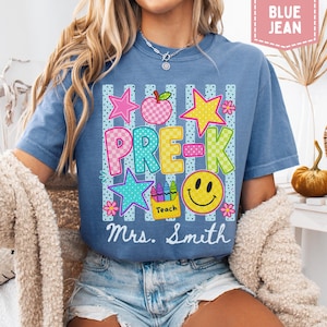 Pode incluir: Uma t-shirt azul jeans com uma estampa gr&aacute;fica contendo o texto PRE-K e Mrs. Smith. O design inclui uma ma&ccedil;&atilde; xadrez, estrelas, um rosto sorridente e uma caixa de giz de cera com a etiqueta Teach sobre um fundo de listras verticais.