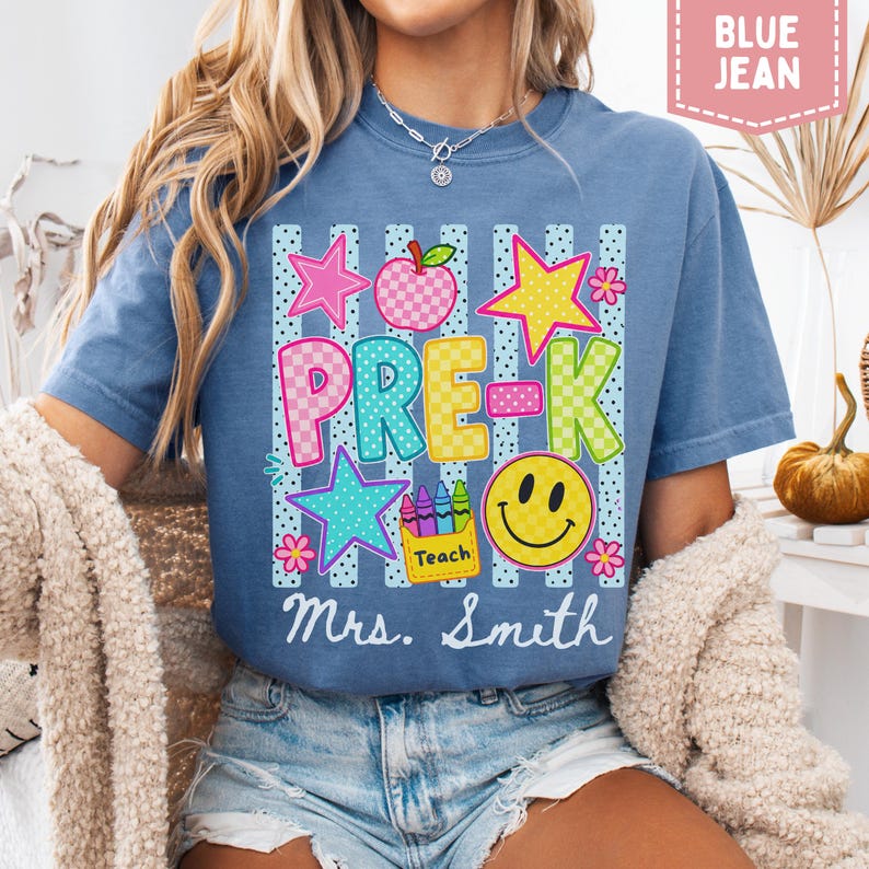Pode incluir: Uma t-shirt azul jeans com uma estampa gr&aacute;fica contendo o texto PRE-K e Mrs. Smith. O design inclui uma ma&ccedil;&atilde; xadrez, estrelas, um rosto sorridente e uma caixa de giz de cera com a etiqueta Teach sobre um fundo de listras verticais.