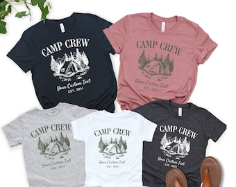 May include: Four t-shirts with a camping theme. The shirts are in different colours: black, grey, white, and pink. Each shirt has a graphic of a tent in a forest with the text "Camp Crew" and "Your Custom Text EST. 2024".